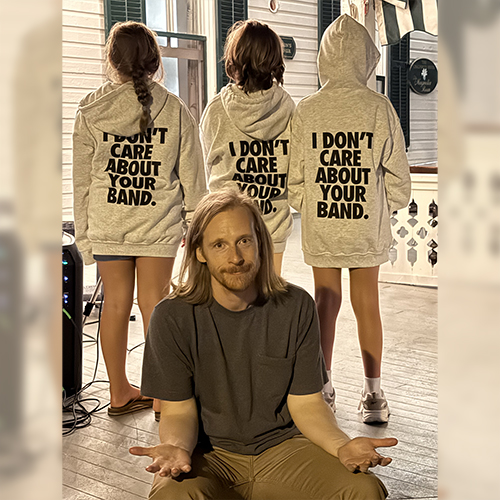 Kevin Devine sitting in front of three kids. The backs of their sweatshirts read I don't care about your band