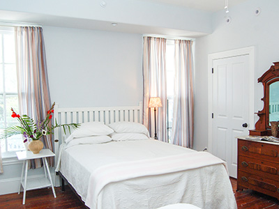 A double bed in a pale blue room with flowers on the nightstand and an antique dresser